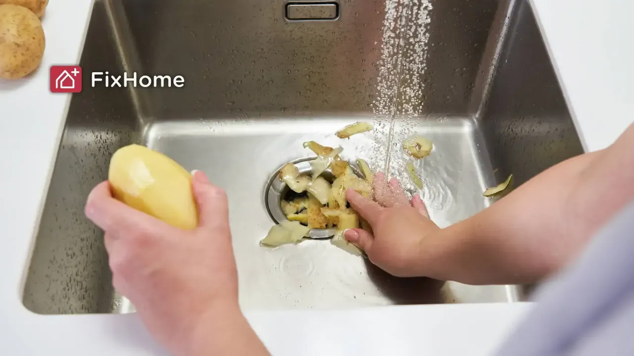 Hands pushing potato peels into a garbage disposal while water runs.