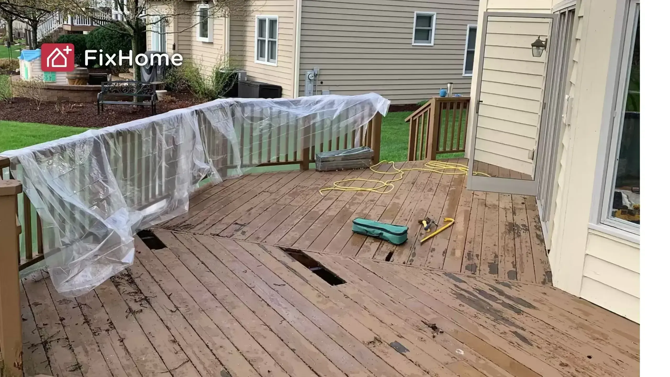 A wooden deck with exposed areas, tools, and plastic covering. Grass and houses in the background
