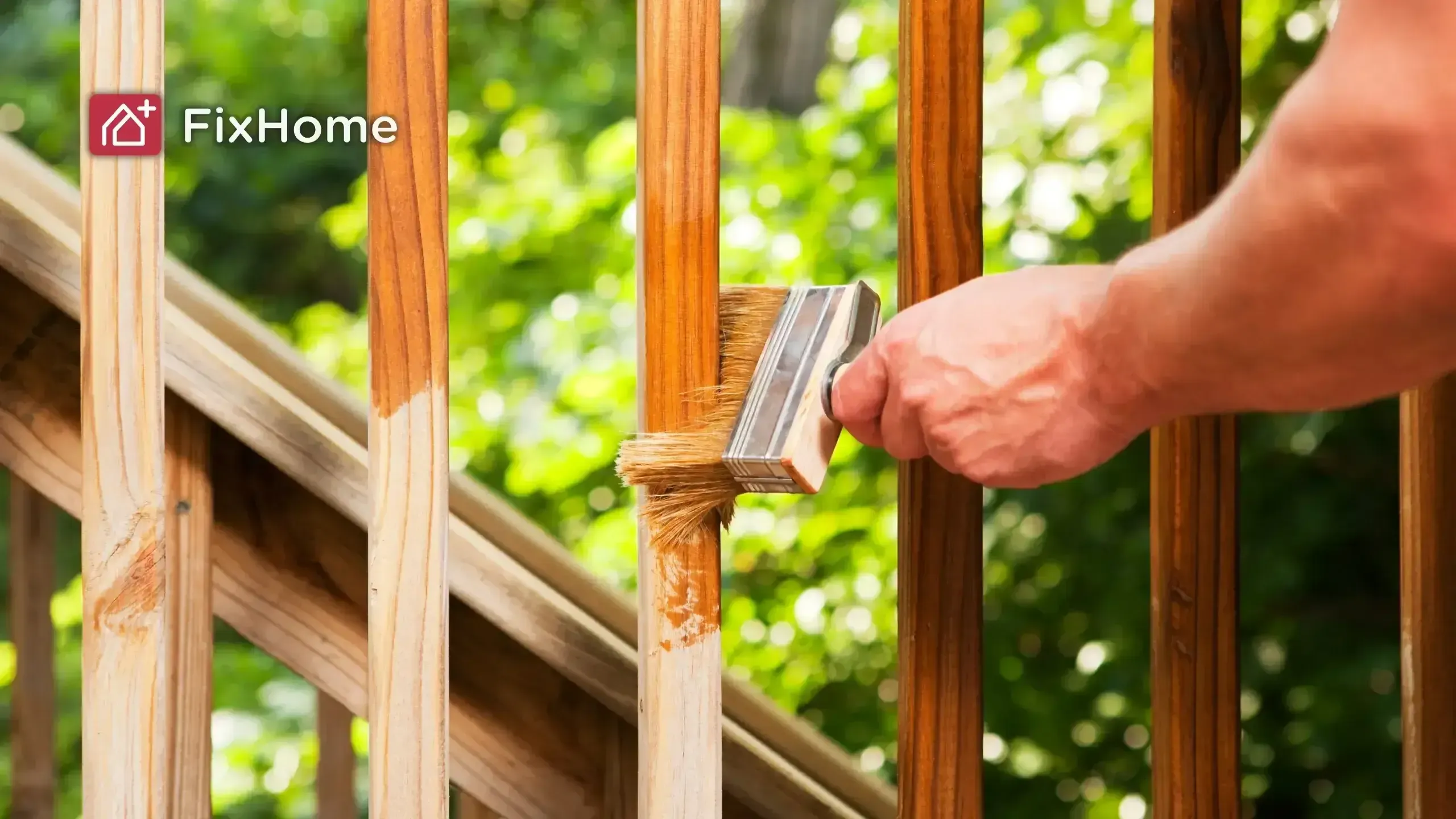 A person stains a wooden deck railing with a brush, surrounded by greenery