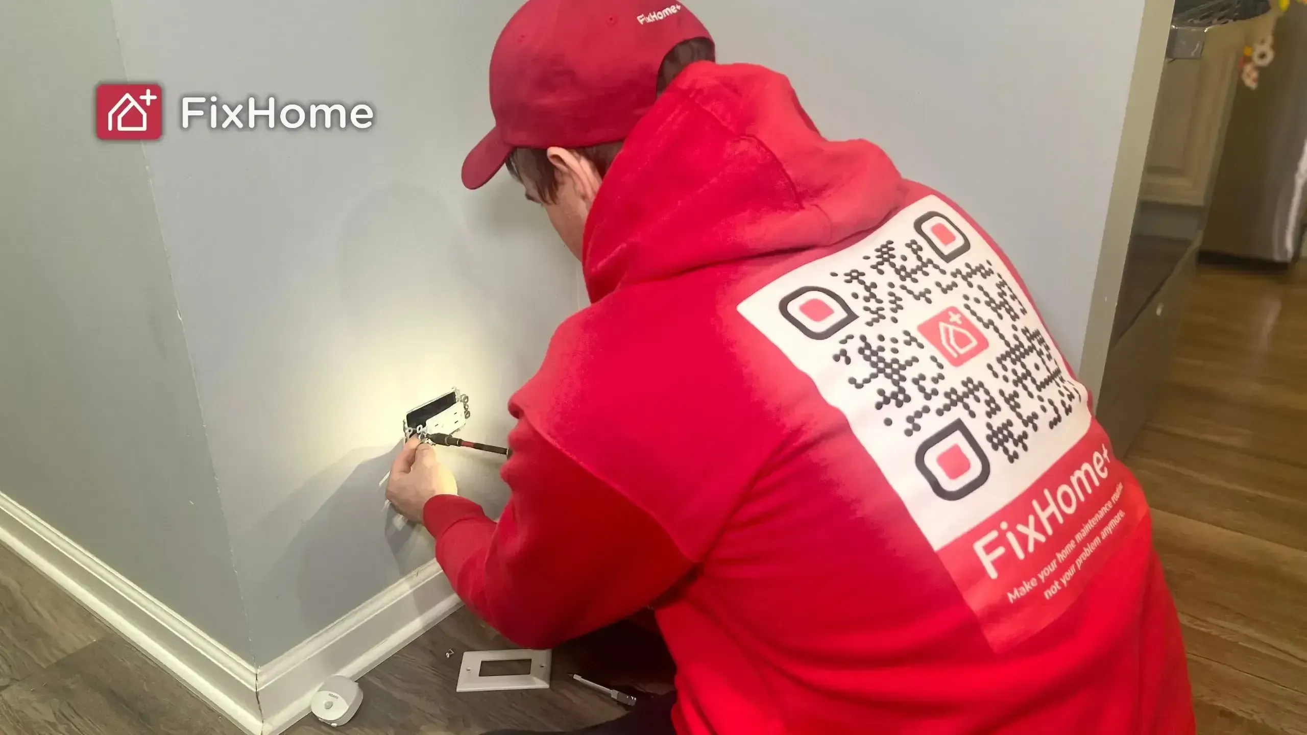 A FixHome handyman in a red hoodie repairing a wall socket in a kitchen, with tools scattered on the floor