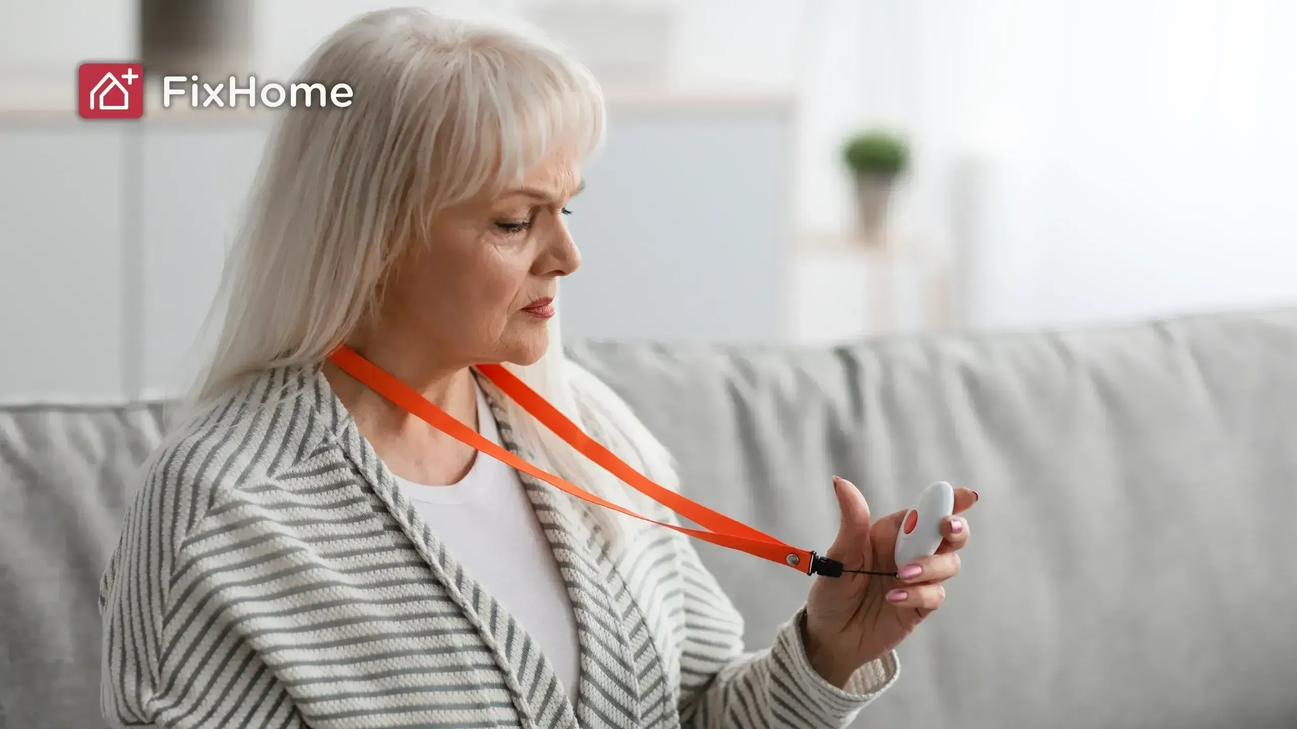 An older woman holds a medical alert device on an orange lanyard while sitting on a couch.