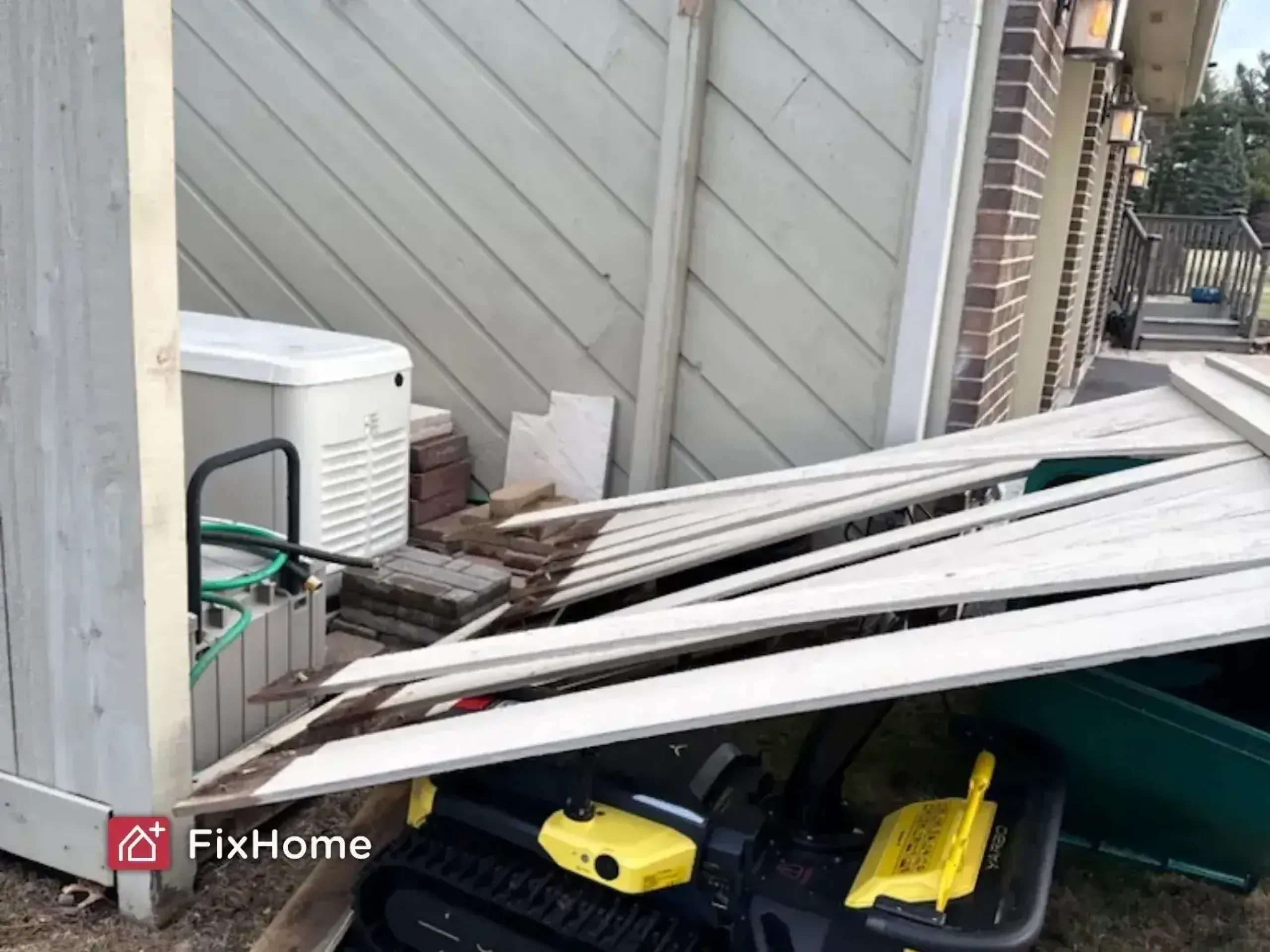 A pile of dislodged wooden planks from a storm-damaged fence is next to a green storage container and an air conditioning unit