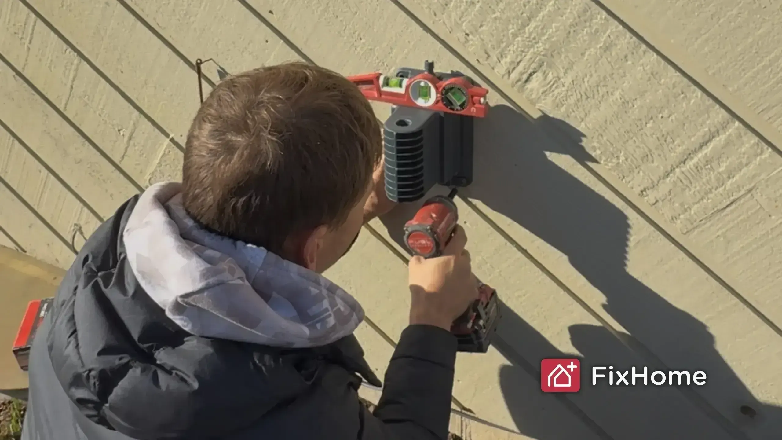 A handyman in a jacket uses a power drill to install a hose reel mount on a beige wall. A red level tool rests above it. The FixHome+ logo is in the corner