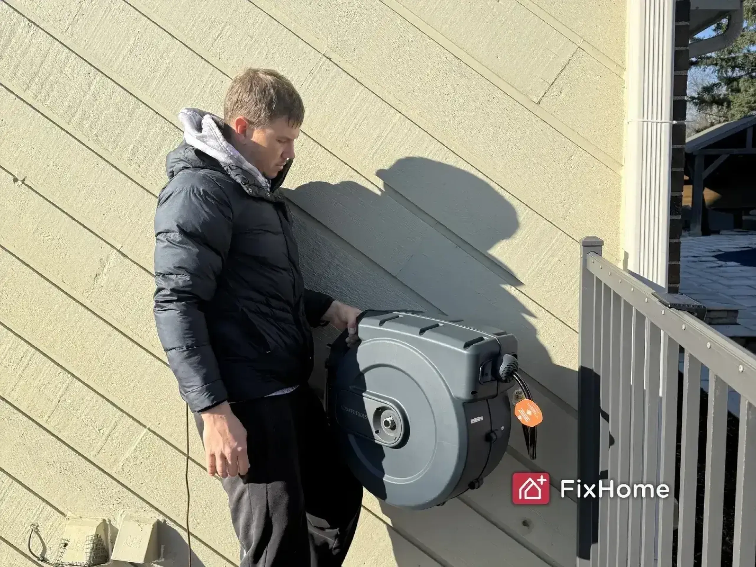 A FixHome+ handyman in a black jacket installs a retractable hose reel on a diagonal wooden wall outdoors, near a metal railing