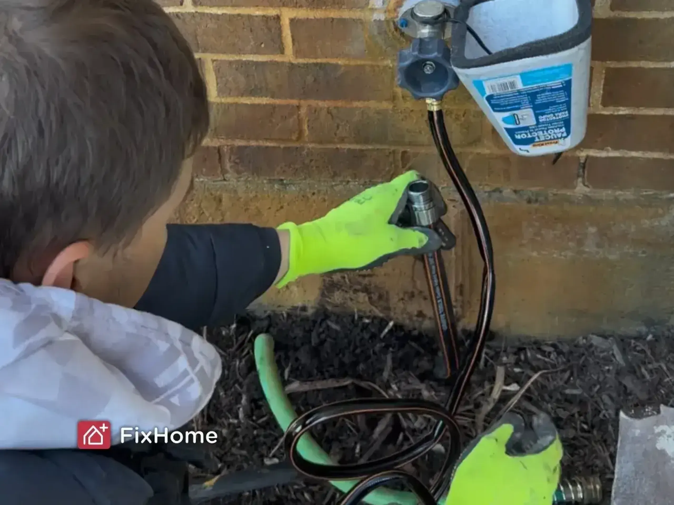 A handyman wearing bright green gloves adjusts an outdoor faucet on a brick wall. Coiled hoses and protective foam are visible, indicating install work.