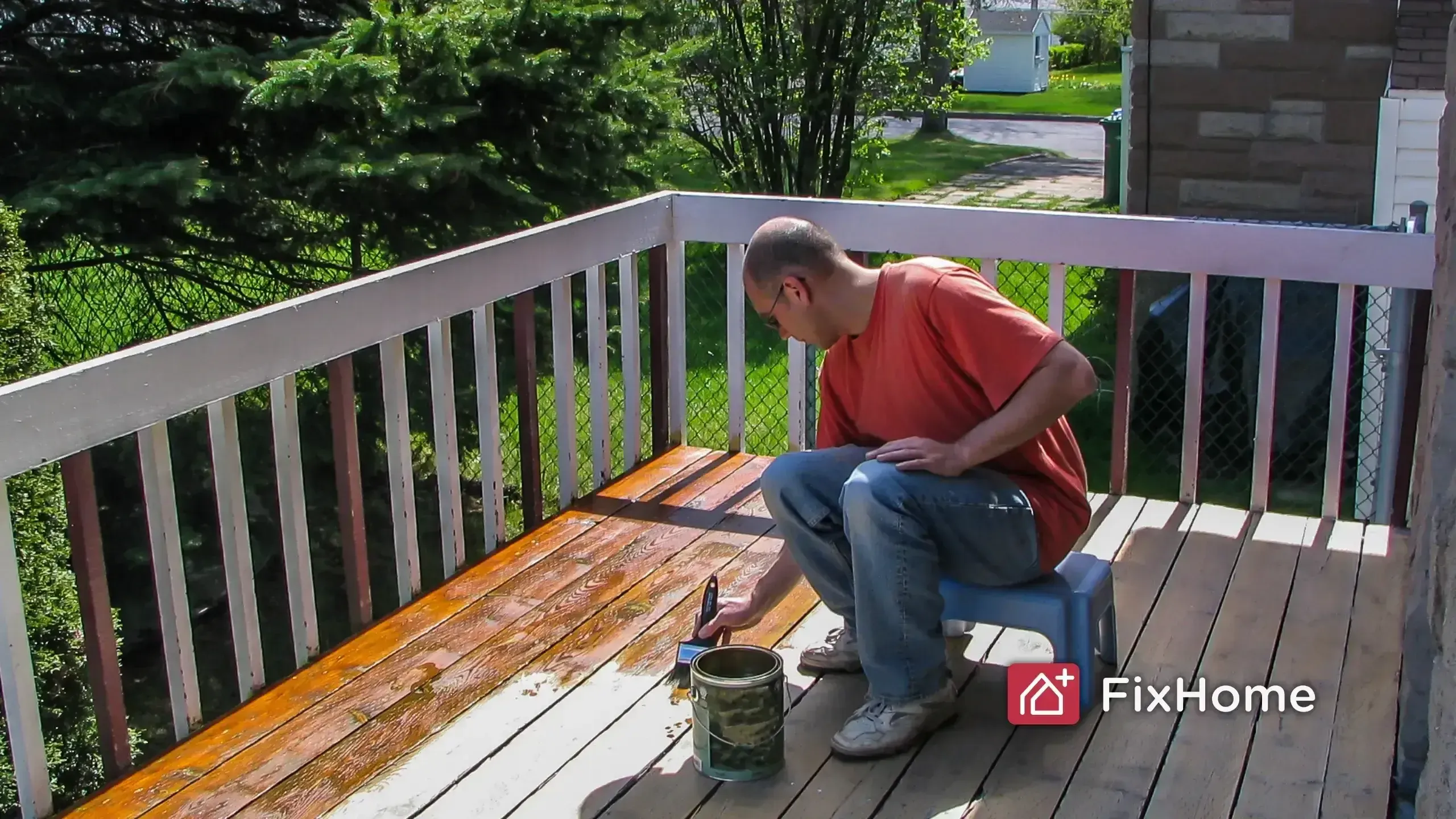 A homeowner sitting on a suburban deck applying stain by hand with a brush and can
