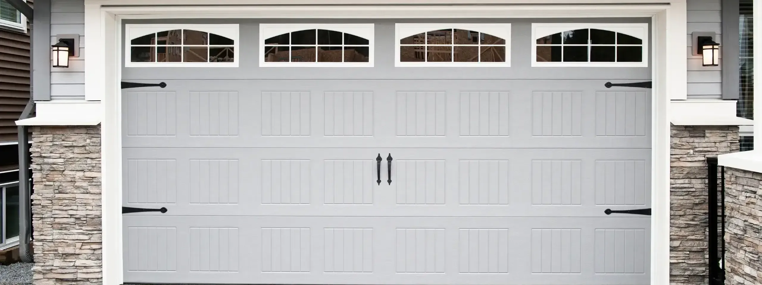 A grey garage door with decorative windows and black handles, next to stone walls. 