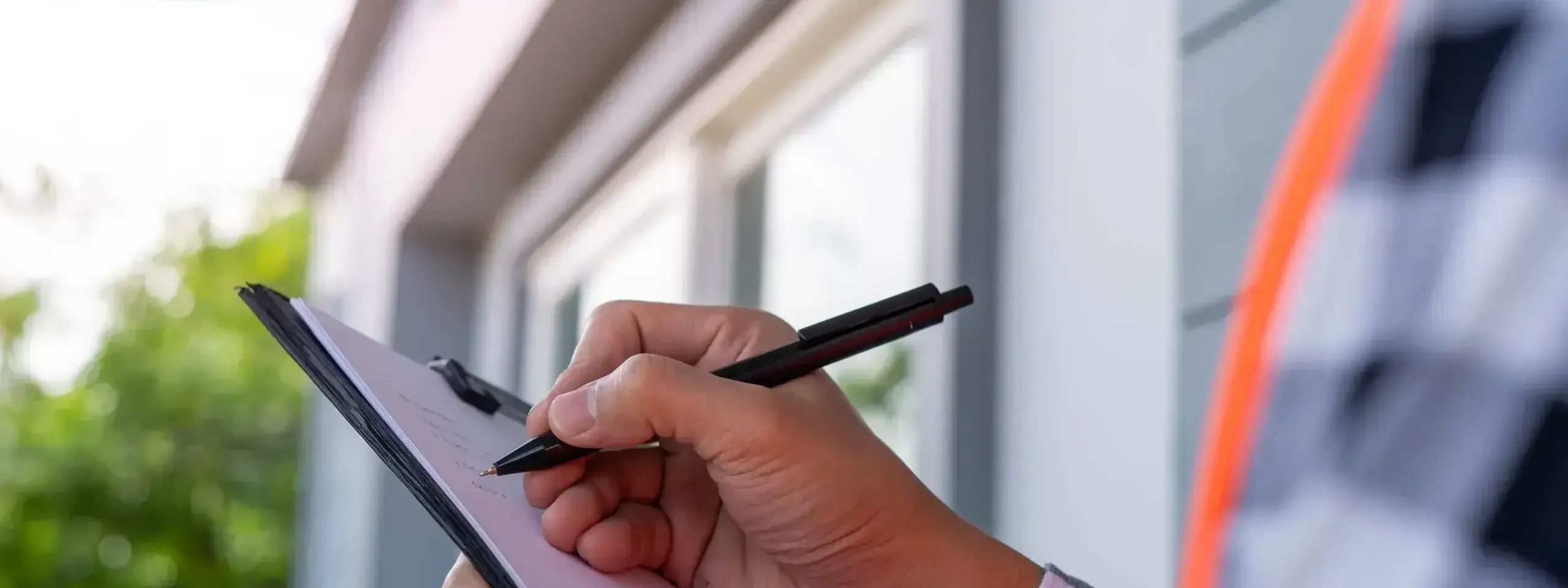A home inspector writing on a clipboard with a pen outside a house, greenery in background. 