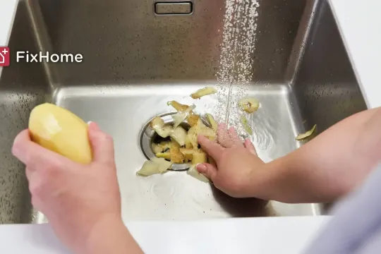 Hands pushing potato peels into a garbage disposal while water runs.