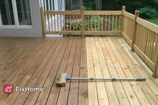 A suburban wood deck mid-staining — half the boards already stained to a warm brown, the other half still bare, with a long-handled brush resting across the work line