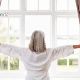 An old woman with silver hair opens the curtains in a bright room